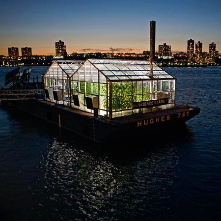 a glass greenhouse with green plants inside floats on a barge at night with the sunset and nyc in the background