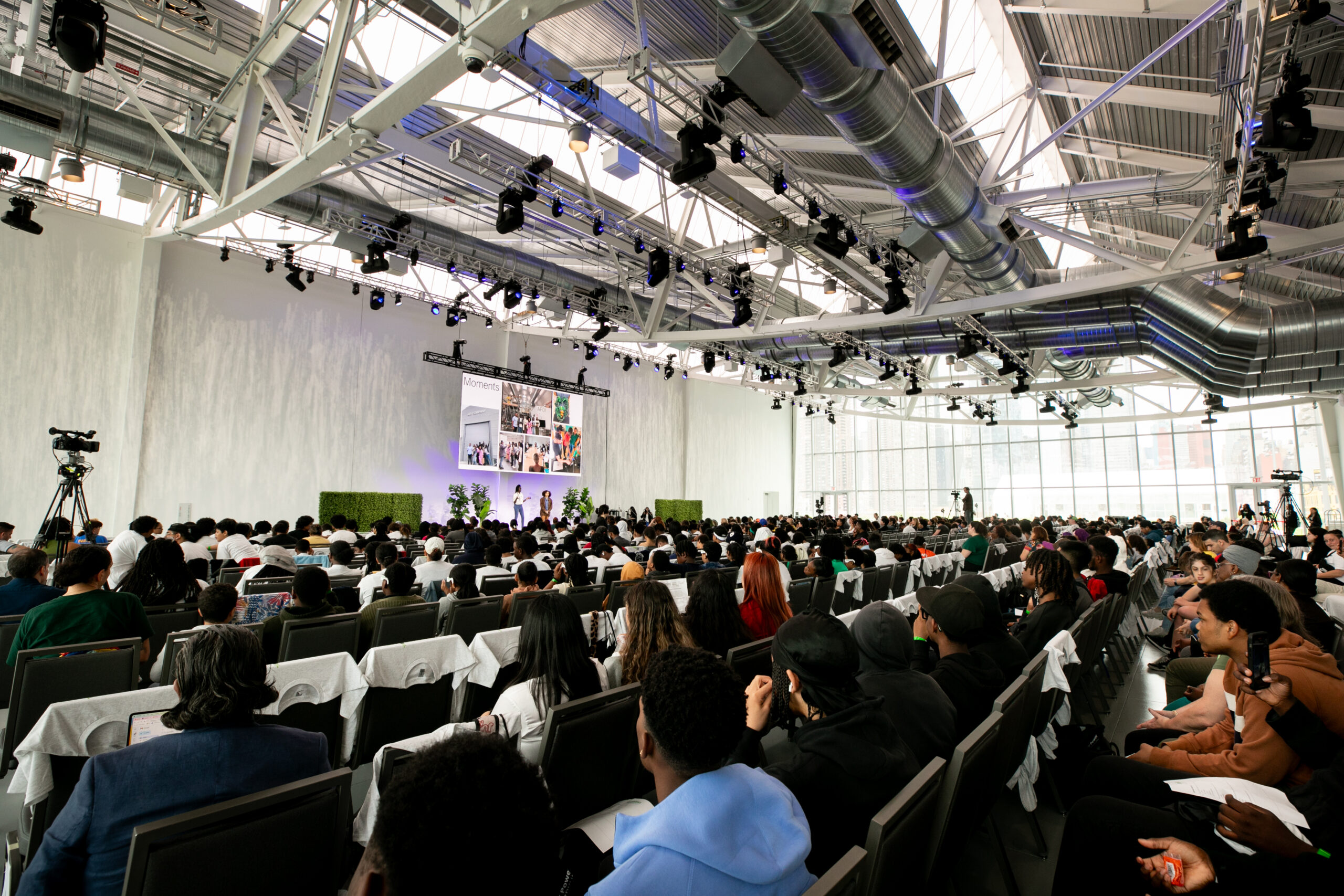 rows of seated students all facing a stage inside a white room with large glass walls located atop the javits center in new york city